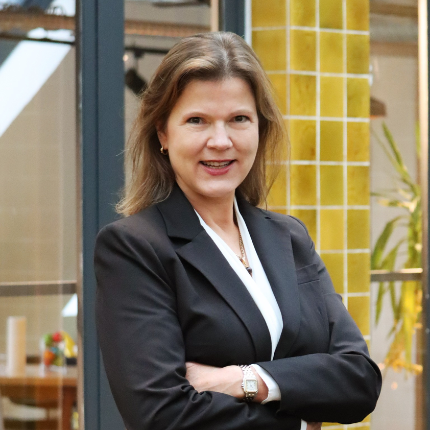 Professionelle Frau in schwarzem Anzug mit verschränkten Armen, lächelnd vor einem modernen Hintergrund mit gelben Fliesen und Pflanzen. Ideal für Themen wie Business, Führung oder Networking.