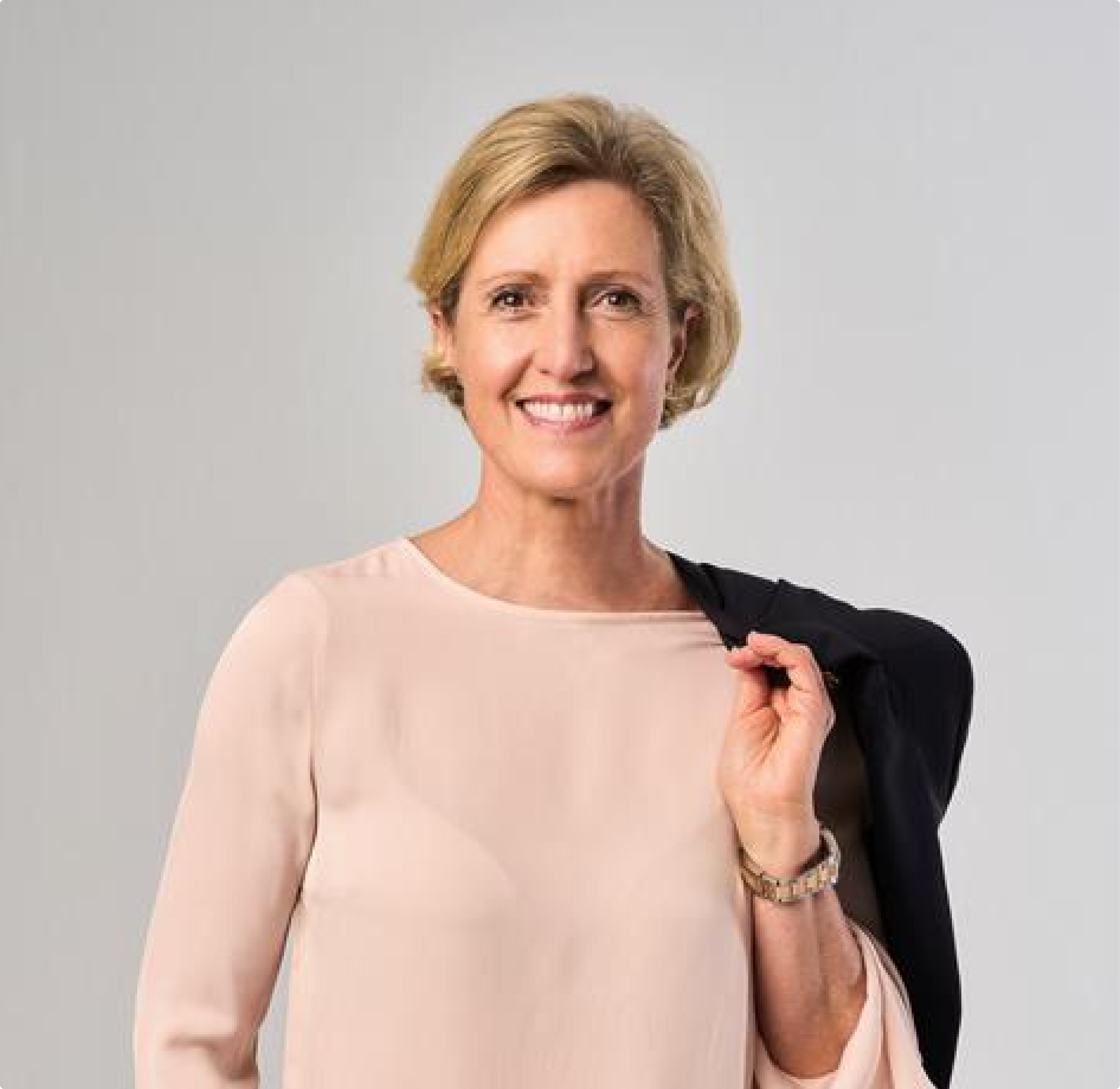 Portrait einer lächelnden, mittelalten Frau in formeller Kleidung, die einen Blazer über der Schulter trägt. Der Hintergrund zeigt eine moderne Bürofläche mit großen Fenstern und natürlichem Licht. Die Frau strahlt Selbstbewusstsein und Professionalität aus.